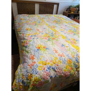 Könnte beinhalten: Eine florale Steppdecke mit einem hölzernen Kopfteil. Die Steppdecke zeigt ein Muster aus bunten Blumen in Rosa-, Gelb-, Blau- und Orangetönen auf weißem Hintergrund. Die Steppdecke ist mit einem Rautenmuster gesteppt.