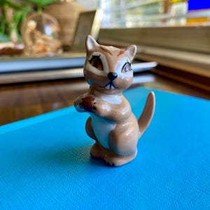 Könnte beinhalten: Kleine Eichhörnchen-Figur aus Keramik in Braun-, Beige- und Weißtönen. Das Eichhörnchen hält einen kleinen, runden Gegenstand, möglicherweise eine Nuss, in seinen Pfoten. Die Figur hat detaillierte Gesichtszüge und steht vor blauem Hintergrund.