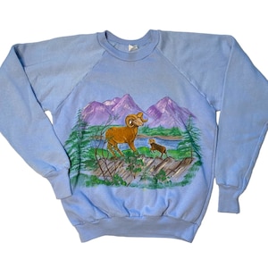 May include: Light blue sweatshirt with a painted design of a bighorn sheep and a lamb in a mountain landscape. The mountains are purple and the sky is blue.