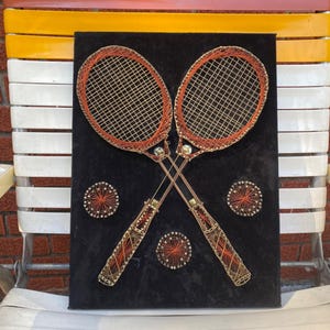 Puede incluir: Una pieza de arte de hilo con temática de tenis que presenta dos raquetas de tenis cruzadas y tres pelotas de tenis. Las raquetas son marrones y doradas, con un fondo negro. Las pelotas de tenis son naranjas y doradas. La obra está montada sobre un lienzo negro.