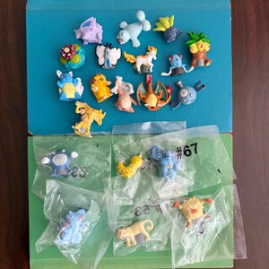 May include: A collection of small, colorful plastic character figurines featuring various creatures, including some loose and others individually sealed in clear plastic bags. Several figures are arranged on a blue surface, while others are on a green surface.
