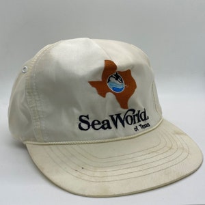 May include: White satin baseball cap with an embroidered logo of SeaWorld of Texas. The logo features a brown outline of the state of Texas with a blue wave and a white whale inside.