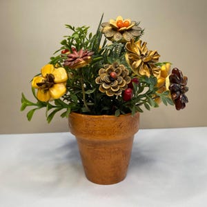 May include: A small terracotta pot filled with a faux floral arrangement. The flowers are made of pinecones and wood and are painted in shades of yellow, brown, and red.