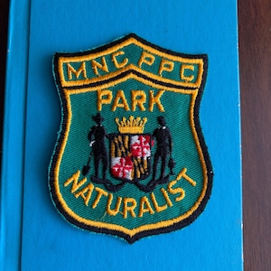 May include: A shield-shaped embroidered patch with a green background and yellow border. It features the text MNC PPC PARK NATURALIST in yellow. The center displays a golden crown above a shield with red, white, and yellow patterns, flanked by two black figures.
