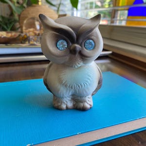 May include: A ceramic owl figurine with a light brown body, dark brown accents, and large blue rhinestone eyes. The owl is perched on a blue book. The figurine has a vintage aesthetic.