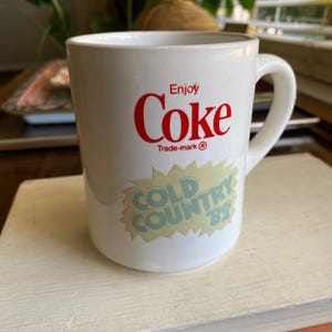 May include: White ceramic mug with the Coca-Cola logo in red, featuring the text "Enjoy Coke" and "Trade-mark ®". A light green and yellow sticker reads "COLD COUNTRY '82". The mug has a curved handle.