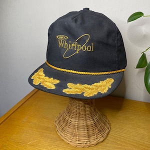May include: A black and gold baseball cap with a gold embroidered Whirlpool logo and a gold rope trim. The cap has a gold embroidered design on the brim.