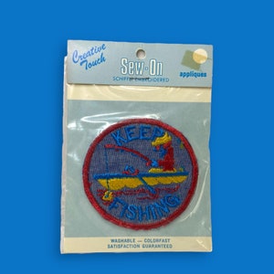 May include: A blue and red embroidered iron-on patch with the text "Keep Fishing" and a cartoon fisherman in a boat. The patch is packaged on a white card with the text "Creative Touch Sew-On appliques Schiffer Embroidered Washable - Colorfast Satisfaction Guaranteed".
