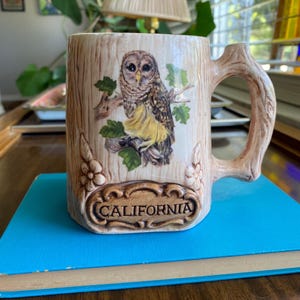 Puede incluir: Una taza de cerámica blanca con un asa negra y el texto "But First, Coffee" en negro, con una fuente de estilo manuscrito. La taza está sobre una superficie blanca.