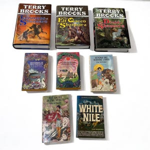 Puede incluir: Una colección de diez libros de bolsillo, incluyendo novelas de fantasía de Terry Brooks, un libro sobre el Nilo Blanco y un libro sobre cenas de pescado en Memison. Los libros tienen portadas coloridas con ilustraciones.