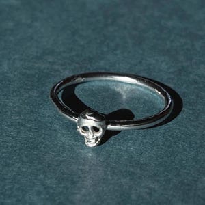 May include: A silver ring with a small skull design. The ring is simple and minimalist.
