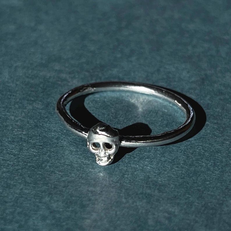 May include: A silver ring with a small skull design. The ring is simple and minimalist.