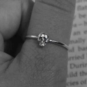 May include: A silver ring with a small skull charm. The ring is worn on a finger.
