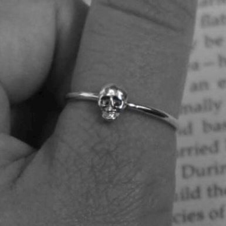 May include: A silver ring with a small skull charm. The ring is worn on a finger.