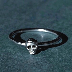 May include: A silver ring with a small skull design. The ring is simple and minimalist, with a smooth band and a detailed skull charm.