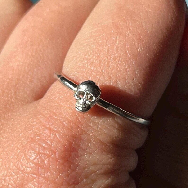 May include: A silver ring with a small skull charm. The ring is thin and delicate.