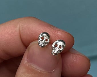 May include: A pair of silver skull stud earrings. Each earring features a detailed skull design with visible eye sockets, nose, and jaw. The earrings are small and appear to be made of a shiny metal.
