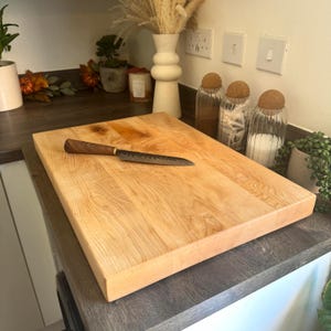 May include: A light-coloured wooden chopping board with a rectangular shape. A small knife with a dark wooden handle and patterned blade is placed on the board. The board is on a kitchen countertop.