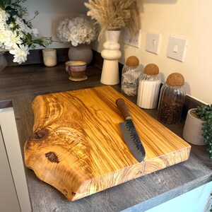 May include: A rectangular wooden cutting board with a natural edge and a dark-handled knife. The board has a light brown color with visible wood grain. The knife is placed on the board, suggesting a culinary theme.