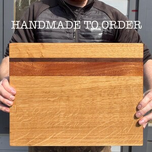 May include: A rectangular wooden cutting board with a light brown oak grain. A strip of darker wood runs horizontally across the center. The text "HANDMADE TO ORDER" is displayed in white letters at the top.