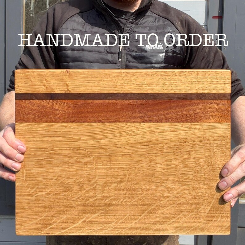 May include: A rectangular wooden cutting board with a light brown oak grain. A strip of darker wood runs horizontally across the center. The text "HANDMADE TO ORDER" is displayed in white letters at the top.
