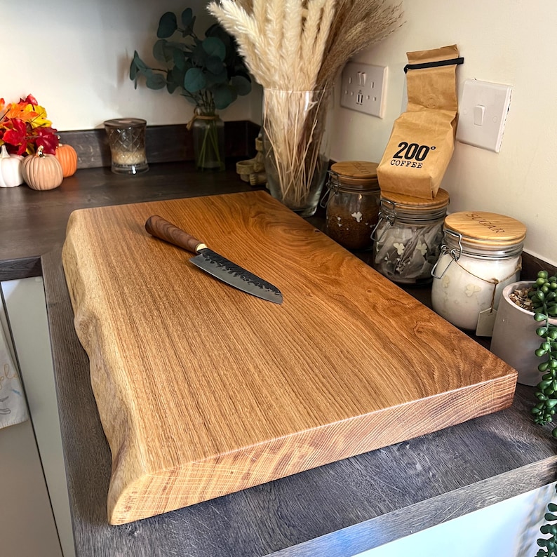 May include: A wooden chopping board with a natural edge and a knife with a dark handle. The board is light brown and rests on a dark countertop. The knife has a silver blade.