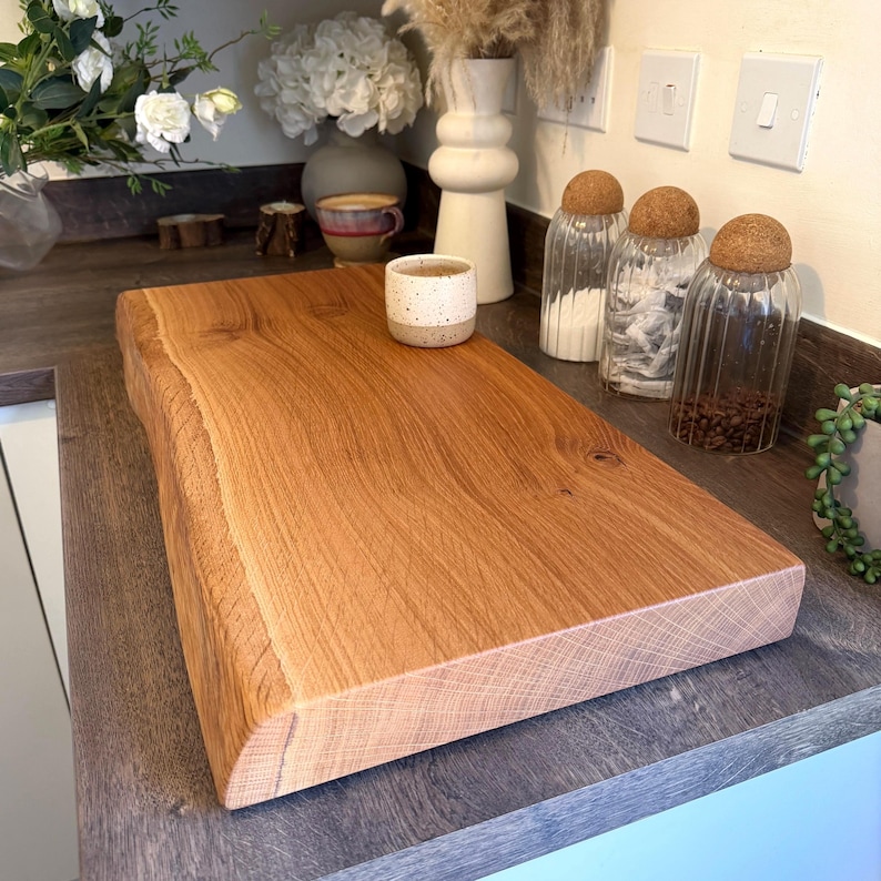 May include: A rectangular, light brown wooden chopping board with a live edge. The board is made of solid wood and has a natural grain pattern. A small, speckled ceramic cup sits on the board. The board is approximately 46 cm long.