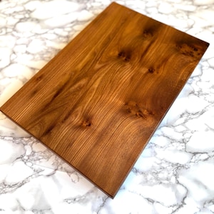 May include: A rectangular wooden cutting board with a rich, warm brown finish. The wood grain is visible, and the board has a smooth surface. The cutting board is set against a white and gray marble background.