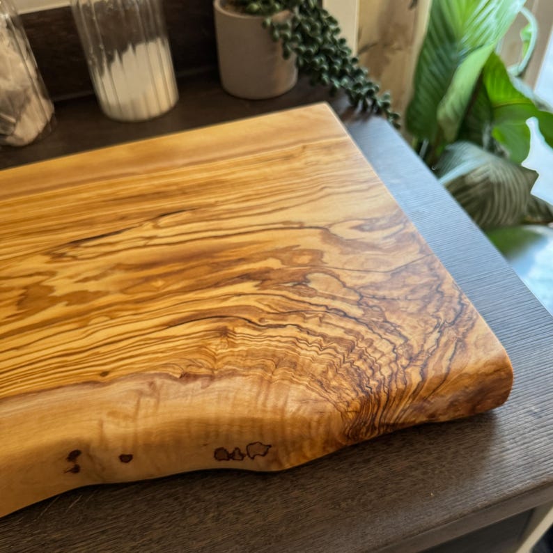 May include: A rectangular wooden cutting board with a natural edge. The board is made of light brown wood with dark brown grain patterns. The board is on a dark brown surface.