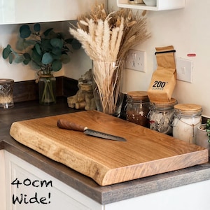 May include: A wooden chopping board, approximately 40cm wide, with a natural edge. A small knife with a wooden handle rests on the board. The board is a light brown colour and sits on a countertop.