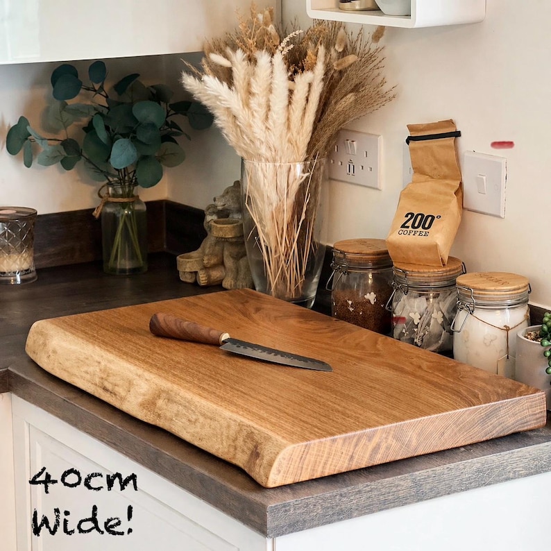 May include: A wooden chopping board, approximately 40cm wide, with a natural edge. A small knife with a wooden handle rests on the board. The board is a light brown colour and sits on a countertop.