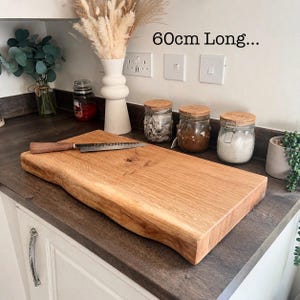 May include: A light brown wooden chopping board, approximately 60cm long, with a natural edge and a knife. The board is on a dark countertop. The text "60cm Long..." is visible.