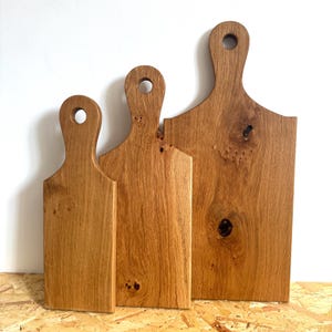 May include: Three wooden cutting boards of varying sizes. Each board is made of light brown wood with a natural grain pattern. They have a handle with a circular hole for hanging. The boards are arranged from smallest to largest.