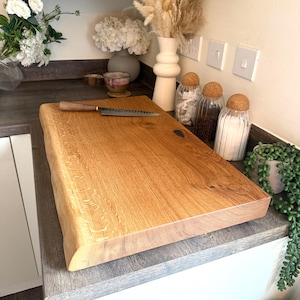 May include: A large, rectangular wooden chopping board with a natural edge and a knife on top. The board is made of light-coloured wood and has a smooth surface. The knife has a wooden handle and a silver blade. Kitchenware.