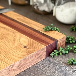 May include: A rectangular wooden cutting board with a light brown oak grain and a dark brown stripe down the center. The end of the board has a dark brown accent. Small green decorative plants are scattered around the board.