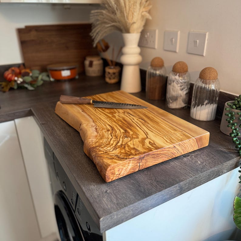 May include: A rectangular wooden cutting board with a natural edge and a dark-handled knife. The board is light brown with a prominent wood grain pattern. The knife is placed on the board.