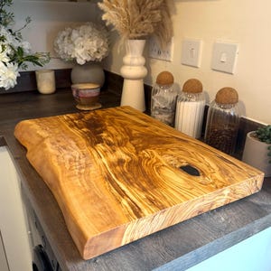 May include: A large, rectangular wooden cutting board with a live edge. The board has a natural wood grain pattern in shades of brown and gold. The board is approximately 18 inches long and 12 inches wide, with a dark knot in the center.