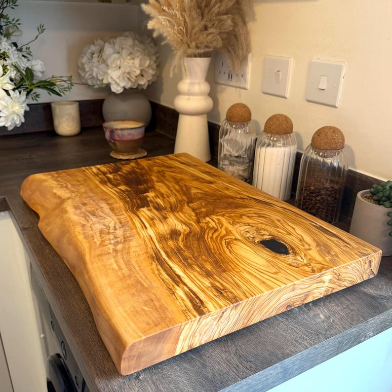 May include: A large, rectangular wooden cutting board with a live edge. The board has a natural wood grain pattern in shades of brown and gold. The board is approximately 18 inches long and 12 inches wide, with a dark knot in the center.