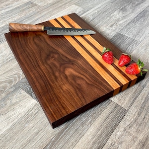 May include: A large, rectangular walnut cutting board with three stripes of lighter wood. The board has a natural wood finish and is shown with a knife and strawberries.