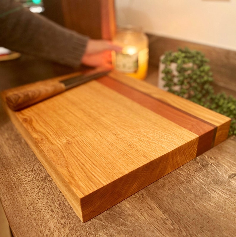 May include: A rectangular wooden cutting board with a light brown oak grain. A strip of reddish-brown wood accents the top edge. A wooden-handled knife rests on the board. The board is approximately 12 inches long and 9 inches wide.