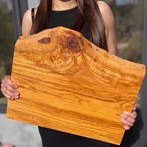 May include: A large, rectangular wooden cutting board with a natural, organic edge. The board is made of olive wood, displaying a warm, golden-brown color with intricate grain patterns. The board is held by a person.