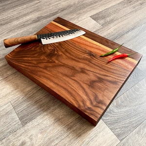 May include: A large rectangular walnut wood cutting board with a contrasting strip of red wood. A black chef's knife and two red chili peppers are on the board.