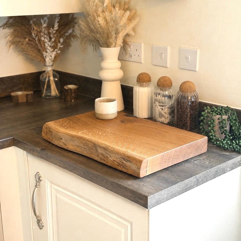 May include: A light brown wooden chopping board with a natural edge is displayed on a kitchen countertop. The board shows the wood grain. A small ceramic cup and glass jars are also visible. The kitchen setting includes a vase with dried flowers.