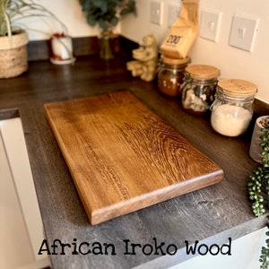 May include: A large, rectangular cutting board made of African Iroko wood. The board has a rich brown color and a natural wood grain pattern. It is sitting on a dark gray countertop.