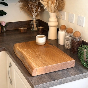 May include: A rectangular, live-edge wooden chopping board with a natural, light brown finish. A small, speckled ceramic cup sits on the board. The board is on a countertop with decorative jars and a vase.