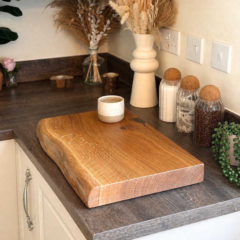 May include: A rectangular, live-edge wooden chopping board with a natural, light brown finish. A small, speckled ceramic cup sits on the board. The board is on a countertop with decorative jars and a vase.