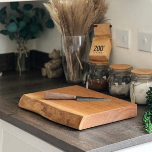 May include: A rectangular wooden chopping board with a live edge, and a small knife with a wooden handle. The board is light brown and sits on a dark countertop. The knife is approximately 20 cm long.