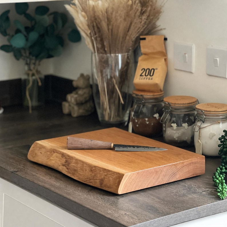 May include: A rectangular wooden chopping board with a live edge, and a small knife with a wooden handle. The board is light brown and sits on a dark countertop. The knife is approximately 20 cm long.