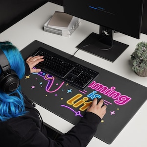 May include: A rectangular black desk mat featuring a neon-style graphic print with the text "gaming is lit" in pink and yellow, accompanied by a stylized game controller outline and small star icons, positioned under a black computer keyboard.