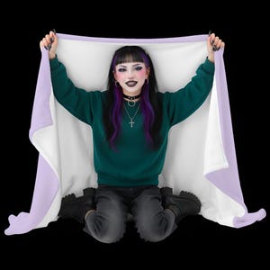 Pode incluir: Um cobertor branco com uma borda lavanda é levantado por uma pessoa vestindo um moletom verde escuro e botas pretas. O cobertor é exibido em um fundo preto. A pessoa está usando um colar gargantilha preto e um colar de cruz de prata.